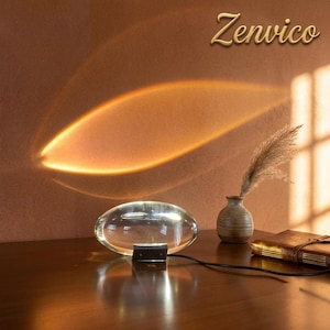 Può includere: Una lampada in vetro trasparente di forma ovale con una base nera e un cavo di alimentazione nero. La lampada proietta un grande fascio di luce dorata ellittica sulla parete. Il testo Zenvico è visibile nell'angolo in alto a destra.
