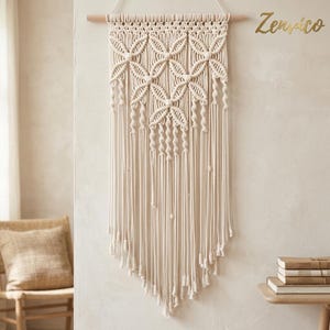Peut inclure: Une tenture murale en macramé de couleur crème suspendue à une tige en bois clair. Le textile présente des motifs noués complexes formant des formes géométriques florales, avec de longues franges pendantes terminées par des nœuds.