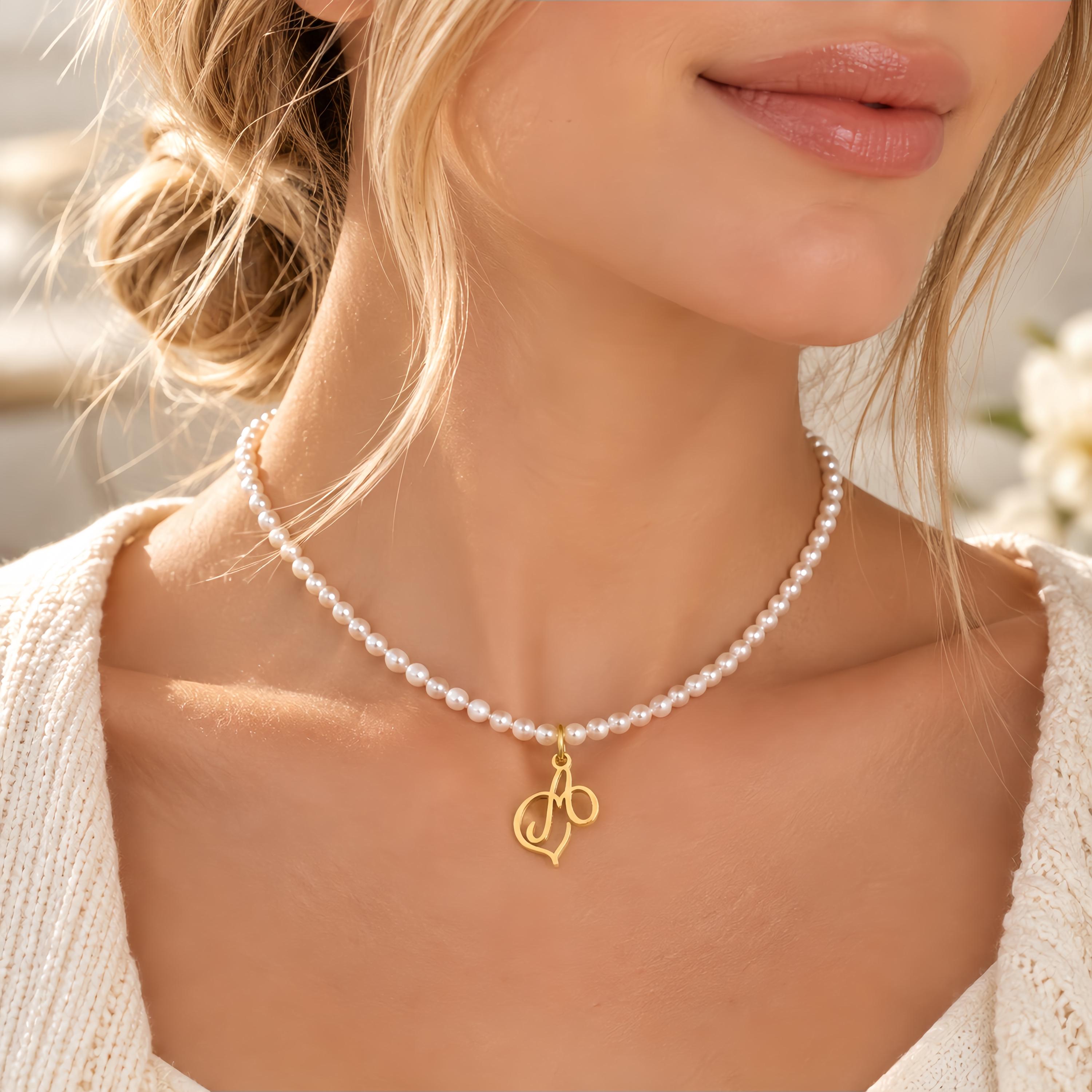 May include: A necklace featuring a strand of small, round white pearls and a gold-toned metal pendant shaped like the cursive letter M. The pendant is suspended from the centre of the pearl strand, which rests around the base of the neck.