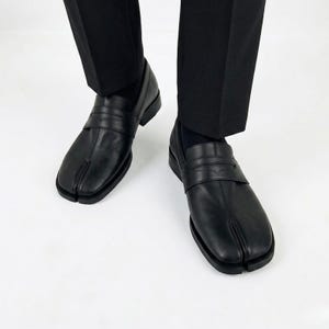 May include: A pair of black leather loafers featuring a distinct split-toe design at the front. The shoes have a classic penny loafer strap across the vamp, a smooth matte finish, and a low heel, worn with black trousers and black socks.