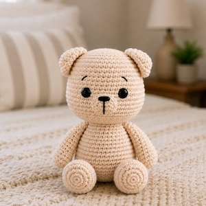 May include: A beige crochet teddy bear with a round head, small ears, and a stout body. The toy features two black plastic eyes, a stitched black nose, and a vertical line for the mouth. The texture is consistent throughout, with visible yarn stitches.