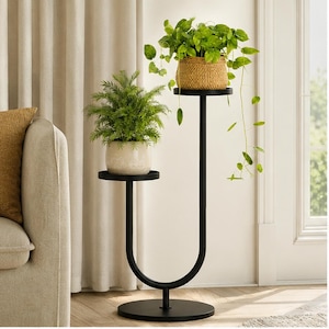 May include: A black metal plant stand with a U-shaped frame and two circular tiers at different heights. The lower tier holds a potted fern in a light-colored ceramic pot, while the higher tier holds a trailing green plant in a woven basket.