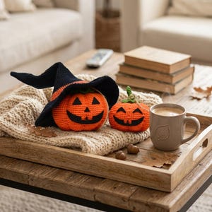 May include: Two crocheted orange pumpkins with black felt jack-o'-lantern faces. The larger pumpkin wears a black crocheted witch hat. They sit on a cream-colored knit fabric next to a light brown ceramic mug with a leaf emblem and two small acorns.