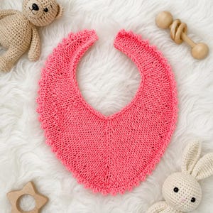 May include: A bright pink knitted baby bib with a scalloped edge, accompanied by a tan crocheted teddy bear, a wooden rattle, a wooden star-shaped teether, and a cream-colored crocheted rabbit toy with a wooden ring base.