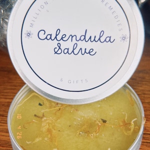 May include: A round silver metal tin containing a yellow-toned salve with visible dried plant pieces. The lid features a white label with the text "MILLION BONTANICAL REMEDIES" and "Calendula Salve & GIFTS" printed in dark blue ink with small flower icons.