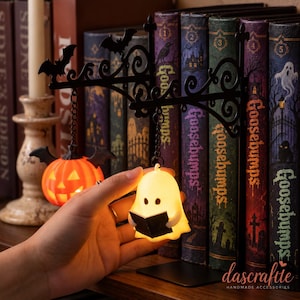 Può includere: Un fermalibri in metallo nero con una staffa decorata a volute, una lanterna a zucca arancione sospesa e una figura di fantasma bianco luminoso che tiene in mano un piccolo libro nero. Il fantasma è illuminato e la lanterna pende da una catena.