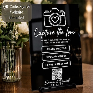 May include: A black rectangular acrylic sign with white text and graphics. It features a camera icon with a heart, the text Capture the Love, Share Photos, Upload Videos, Leave a Message, a QR code, and the names Emma & Daniel with 04.12.26.