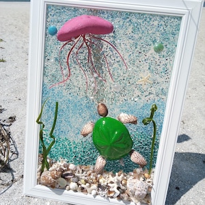 May include: A framed mixed media wall art piece featuring a pink jellyfish and a green sea turtle made of shells and stones. The background is composed of crushed blue glass, with various seashells and white coral pieces at the bottom.