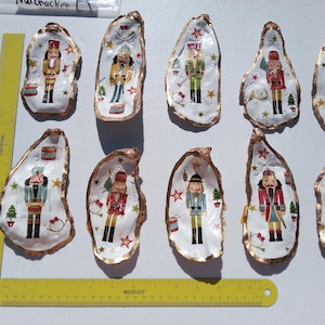 Peut inclure: Collection de coquilles d'huîtres peintes à la main avec différents motifs de soldats Casse-Noisette. Chaque coquille a un intérieur blanc avec un bord doré et est décorée d'étoiles festives et de motifs de sapins de Noël. Une règle jaune est présente.