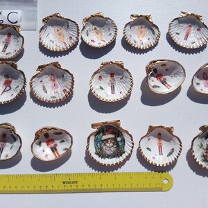 May include: A selection of painted scallop shells with gold edging. Each shell displays a different Christmas design, such as a snowman, a cat in a Santa hat, and a toy soldier. The shells are arranged on a white surface.
