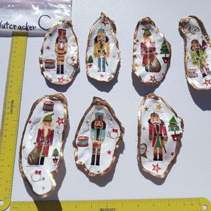 May include: Hand-painted oyster shells with gold trim, each featuring a Nutcracker soldier design. The shells are decorated with red, green, and gold accents, and a small Christmas tree. A ruler is in the image.