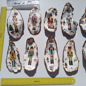 May include: Hand-painted oyster shells with gold trim, each featuring a different Nutcracker soldier design. The shells are white with colorful illustrations of Nutcrackers, Christmas trees, and stars. A yellow ruler is next to the shells.