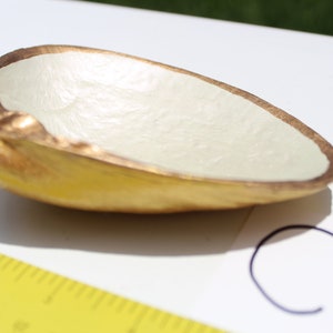 Può includere: Una piccola ciotola bianca con un bordo dorato. La ciotola è realizzata in un materiale liscio e testurizzato e ha la forma di una conchiglia.