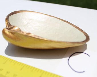 Puede incluir: Un pequeño cuenco blanco con un borde dorado. El cuenco está hecho de un material liso y texturizado y tiene forma de concha.