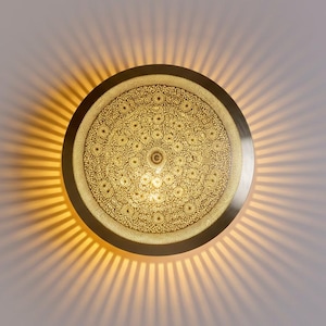 May include: A circular wall lamp with a metallic gold finish and an intricate, perforated floral pattern across its surface. The light fixture emits a warm, glowing radiance that projects a sunburst effect of light rays onto the surrounding area.