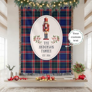 May include: A woven throw blanket featuring a dark blue, green, and red plaid pattern. A large white oval in the center displays a red and gold nutcracker illustration, holly sprigs, and the text THE ANDERSON FAMILY EST. 2026 with fringed edges.