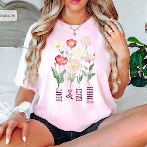 Könnte beinhalten: Ein hellrosa T-Shirt mit einer Grafik aus verschiedenen bunten Blumen in Rot-, Gelb- und Weißtönen. Unter der Blumenillustration steht der Text "ROOT for EACH OTHER" in einer dunkelroten Serifenschrift. Das T-Shirt hat kurze Ärmel.