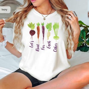 Puede incluir: Una camiseta de color marfil con un gráfico de cinco coloridas hortalizas de raíz, incluyendo una remolacha, una zanahoria, una zanahoria morada, una chirivía y un nabo. Debajo, el texto Let's Root For Each Other en letra morada.