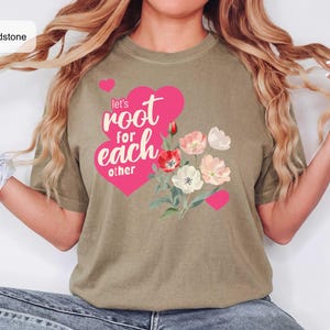 Puede incluir: Una camiseta color arena con un gráfico de un corazón rosa grande y el texto 'let's root for each other' en letras blancas. A la derecha del corazón hay un grupo de flores estilo acuarela en tonos rojos, rosas y blancos.