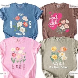 Peut inclure: Quatre t-shirts à manches courtes dans les couleurs bleu jean, espresso, fleur et grès. Chaque t-shirt présente des motifs floraux et le texte "Root for each other" dans diverses dispositions, dont un cœur rose sur le modèle espresso.