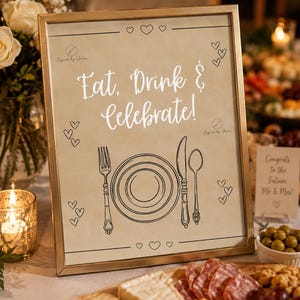May include: A rectangular gold-toned frame containing a beige sign with the text Eat, Drink & Celebrate! in white script. The sign features black line art of a plate, fork, knife, and spoon, surrounded by small heart outlines and two logos.