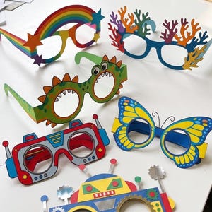 Könnte beinhalten: Eine Sammlung von fünf bunten Papierbrillen mit verschiedenen Motiven, darunter ein Regenbogen mit Sternen, Korallenformen, ein grüner Dinosaurier, ein blau-gelber Schmetterling und ein bunter Roboter mit Antennen und Zahnrad-Details.