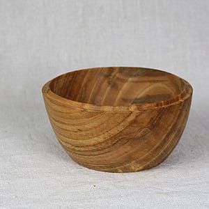 Pode incluir: Uma tigela de madeira redonda com uma cor castanho-claro natural e padrões visíveis de veios de madeira castanho-escuros. A tigela tem um exterior liso e curvo e uma parte superior larga e aberta, exibindo a textura orgânica e os anéis do material.