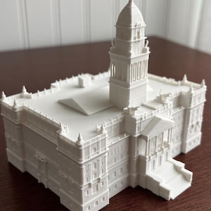 May include: A white 3D printed architectural model of a building featuring a central domed tower, a rectangular main structure with detailed facade elements, corner turrets, and a front entrance with stairs leading up to a portico with columns.