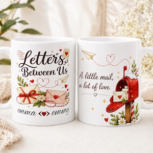 May include: Two white ceramic mugs featuring colorful illustrations of envelopes, a red mailbox, and floral accents. The left mug displays the text Letters Between Us and the names emma and emmy. The right mug features the text A little mail, a lot of love.