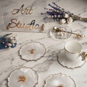 Könnte beinhalten: Ein Set aus drei weißen Untersetzern aus Kunstharz mit gewelltem Rand und goldenen Metallflocken in der Mitte. Eine weiße Keramiktasse mit Goldrand steht auf einem Untersetzer. Ein transparentes Schild zeigt den Text Demet Art Studio in Gold.