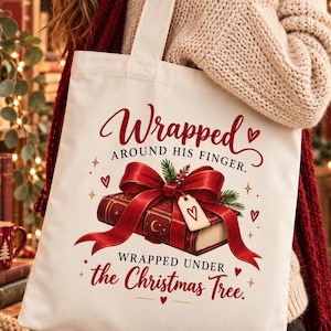 Könnte beinhalten: Eine weiße Stofftasche mit einer Grafik eines roten Buches, das mit einer großen roten Schleife gebunden ist. Der Text lautet Wrapped AROUND HIS FINGER und WRAPPED UNDER the Christmas Tree, verziert mit kleinen roten Herzen und goldenen Sternen.