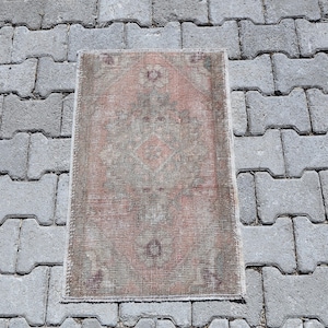 Puede incluir: Una alfombra rectangular con una paleta de colores desvaídos y apagados en rosa viejo, verde salvia y gris carbón. La alfombra presenta un motivo central de medallón de diamante rodeado de intrincados patrones geométricos desgastados y un borde fino deshilachado.