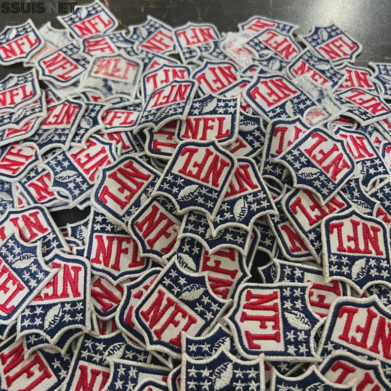 Pode incluir: Uma grande cole&ccedil;&atilde;o de emblemas bordados em forma de escudo com o log&oacute;tipo da NFL em vermelho, branco e azul. Cada emblema apresenta as letras vermelhas NFL sobre um gr&aacute;fico de uma bola de futebol americana branca, rodeada por estrelas.