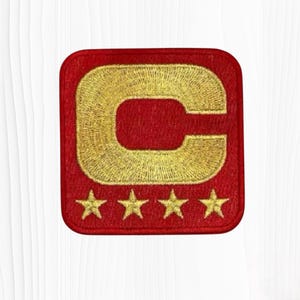 May include: A square-shaped embroidered patch with rounded corners featuring a red background. The design includes a large, bold, gold-colored letter C centered in the upper portion, positioned above a horizontal row of four small gold stars.