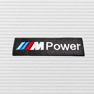May include: A rectangular embroidered patch with a black background featuring the M Power logo. The design includes three slanted stripes in light blue, dark blue, and red on the left side, followed by the white text M Power in a bold font.