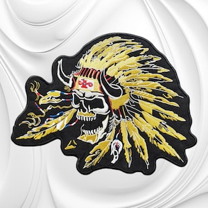 May include: An embroidered patch featuring a skull wearing a large yellow feathered headdress with black horns. The skull is white with black shading, and the headdress includes a central band with a red and white cross symbol and dangling feathers.