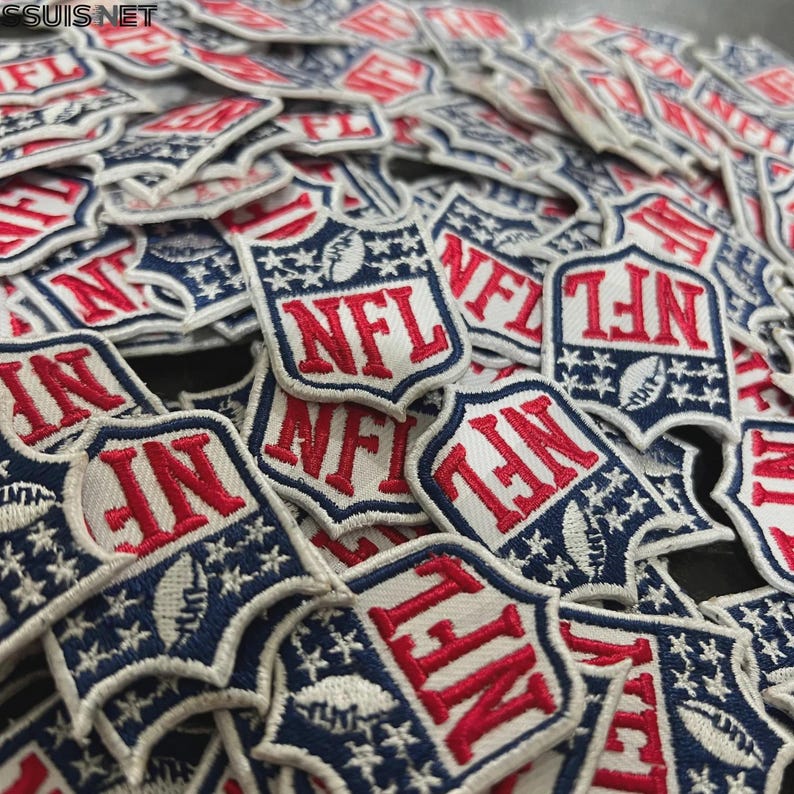Pode incluir: Uma cole&ccedil;&atilde;o de emblemas bordados em forma de escudo com o log&oacute;tipo da NFL em vermelho, branco e azul. Cada emblema apresenta o texto NFL em letras vermelhas, um &iacute;cone de bola de futebol americano branco e estrelas brancas sobre fundo azul.