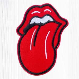 May include: An embroidered patch featuring a stylized red mouth with thick black outlines, white teeth, and a prominent red tongue extending downward. The design is symmetrical with a bold red border and a black inner edge, creating a graphic icon.