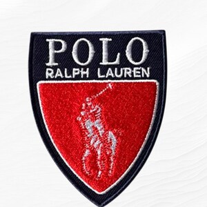 May include: A shield-shaped embroidered patch featuring a black upper section with the white text POLO RALPH LAUREN and a red lower section containing a white embroidered polo player on horseback icon with a mallet raised in the air.
