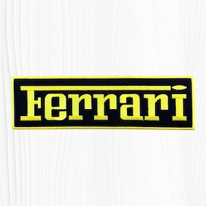 May include: A rectangular embroidered patch featuring a black background with a bright yellow border and the word Ferrari stitched in a bold yellow font across the center. The design includes a horizontal yellow line above the text.