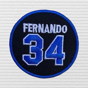 May include: A circular embroidered patch with a black background and a blue border. The patch features the white text "FERNANDO" arched across the top and the large blue number "34" centered below, both outlined in white stitching.