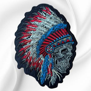 May include: An embroidered patch featuring a profile view of a human skull wearing a large, elaborate headdress. The headdress is composed of numerous feathers in shades of red, white, and blue, with a decorative blue and red band across the forehead.