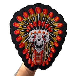 May include: A black embroidered patch featuring a central white skull wearing a vibrant headdress with red and orange circular accents. The skull has red markings around the eyes and a detailed jaw, set against a scalloped black fabric background.