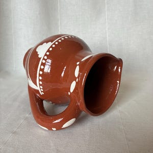 May include: A glossy terracotta-coloured ceramic pitcher featuring a rounded body, a wide spout, and a curved handle. The exterior is decorated with hand-painted white patterns, including dotted lines and floral-inspired shapes, against the dark clay.