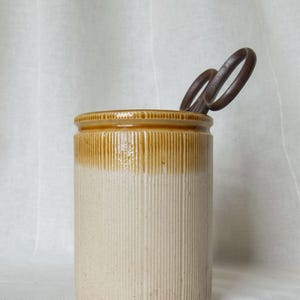 Puede incluir: Un recipiente cilíndrico de cerámica con textura acanalada, que presenta un esmalte brillante de color tostado en la parte superior y un acabado mate de color crema en el cuerpo inferior. Unas tijeras de metal marrón oscuro sobresalen del interior.