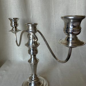 May include: A silver-coloured metal candelabra featuring three candle holders of varying heights. The central holder is elevated, while two curved arms extend outwards to support smaller holders on either side, all set upon a circular base.