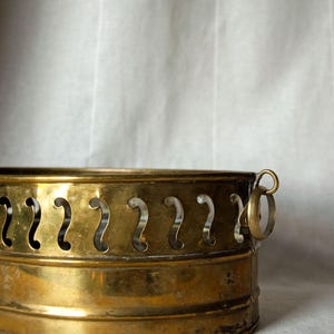 May include: A round brass planter featuring a decorative upper band with a series of vertical, S-shaped cutouts. The vessel has a polished metallic finish and includes a small, circular brass ring handle attached to the side of the rim.