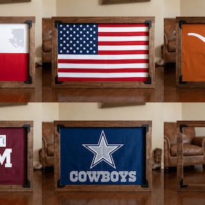 May include: A collection of six rectangular wooden frames, five of which contain fabric displays featuring the Texas flag, the American flag, a white longhorn silhouette on orange, the A&M logo on maroon, and the Cowboys star logo on blue.