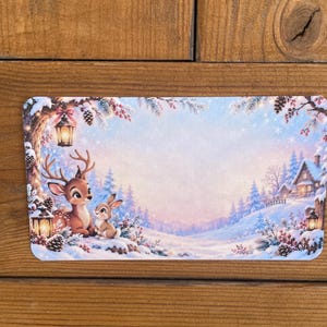 Könnte beinhalten: Eine rechteckige Karte mit einer Winterszene, die einen braunen Hirsch und ein kleines Kaninchen im Schnee zeigt. Die Illustration enthält Tannenbäume, eine leuchtende Laterne an einem Ast und eine kleine Hütte im verschneiten Hintergrund.