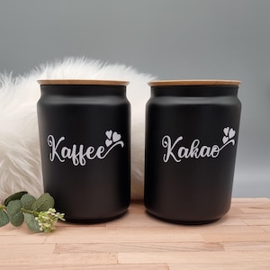 Könnte beinhalten: Zwei mattschwarze, zylindrische Vorratsdosen mit hellen Holzdeckeln. Die linke Dose trägt den weißen Schriftzug "Kaffee" mit drei kleinen Herzen, die rechte Dose den weißen Schriftzug "Kakao" mit drei kleinen Herzen.